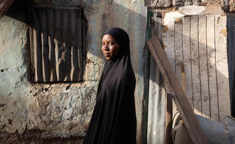 An adolescent girl in a black headscarf stands in front of a courtyard door.