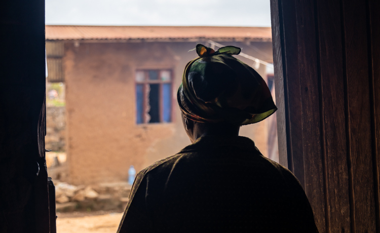 A woman in a head wrap is seen in silhouette peering out a doorway into a sunny courtyard.