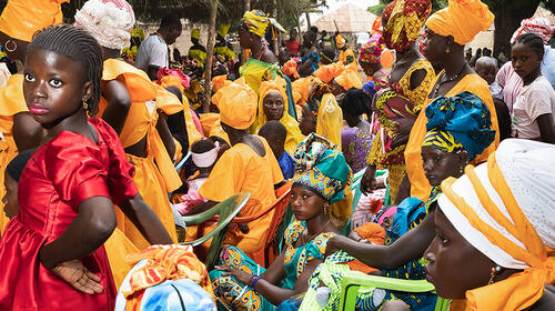 Women and girls gather, sitting and standing in rows, wearing colourful occasion dresses and headwraps.